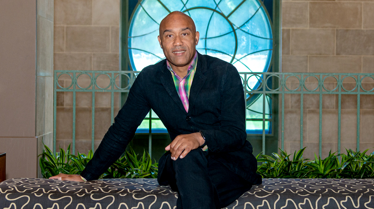 Dr. Augustus Casely-Hayford, wearing a suit, sits on a patterned bench in front of a blue stained-glass window.